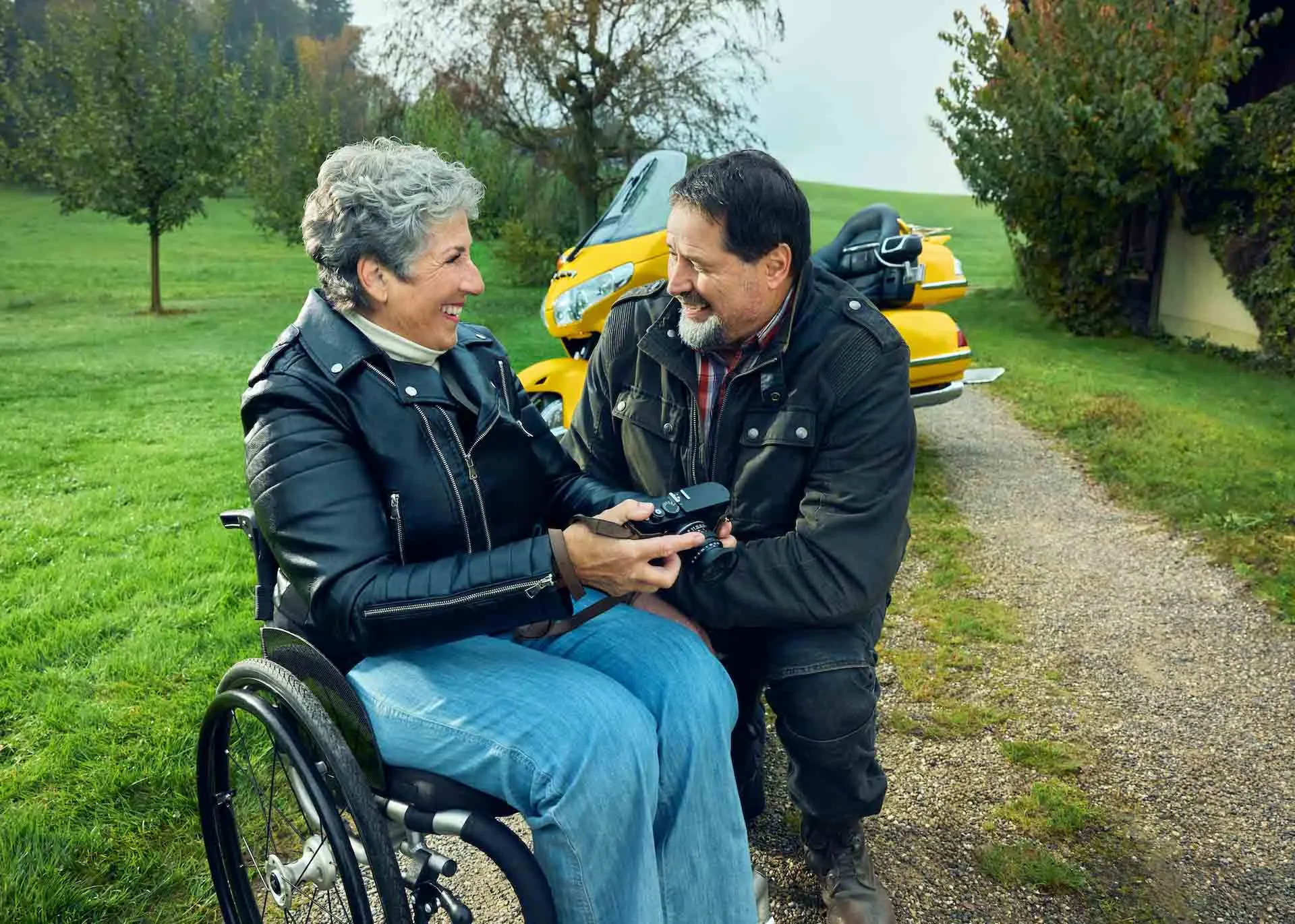 Rosa Zaugg è seduta in carrozzina su un vialetto di ghiaia, tiene in mano una macchina fotografica e sorride al suo partner Lukas.