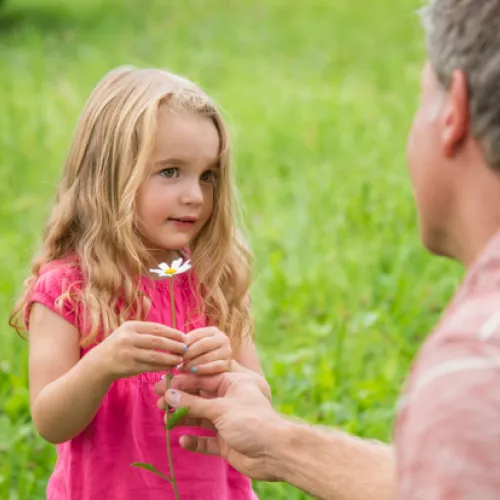 Un père et sa fille dans un pré. Le père offre une fleur à sa fille, qui sourit. 
